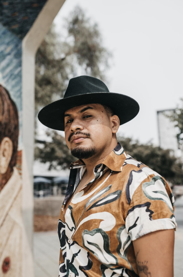 Photo of Yosimar Reyes. Wearing black hat and brown dn white collared shirt. Posing outside.