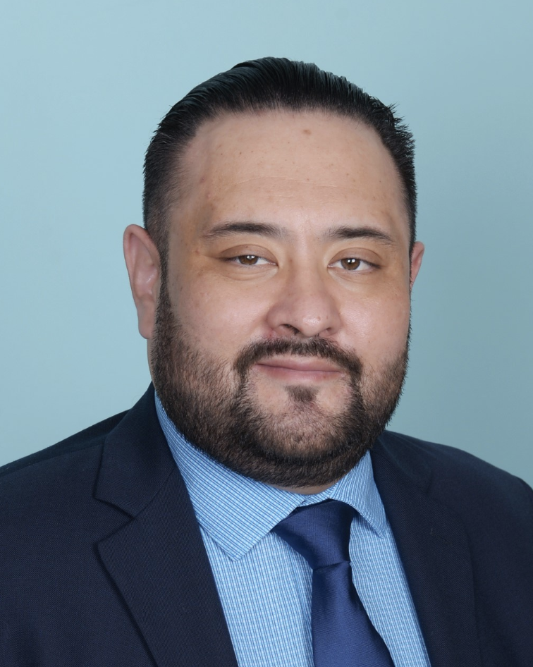 Headshot of Jose wearing a suit and tie. He has fair complexion and a beard with slicked back hair. Blue background.