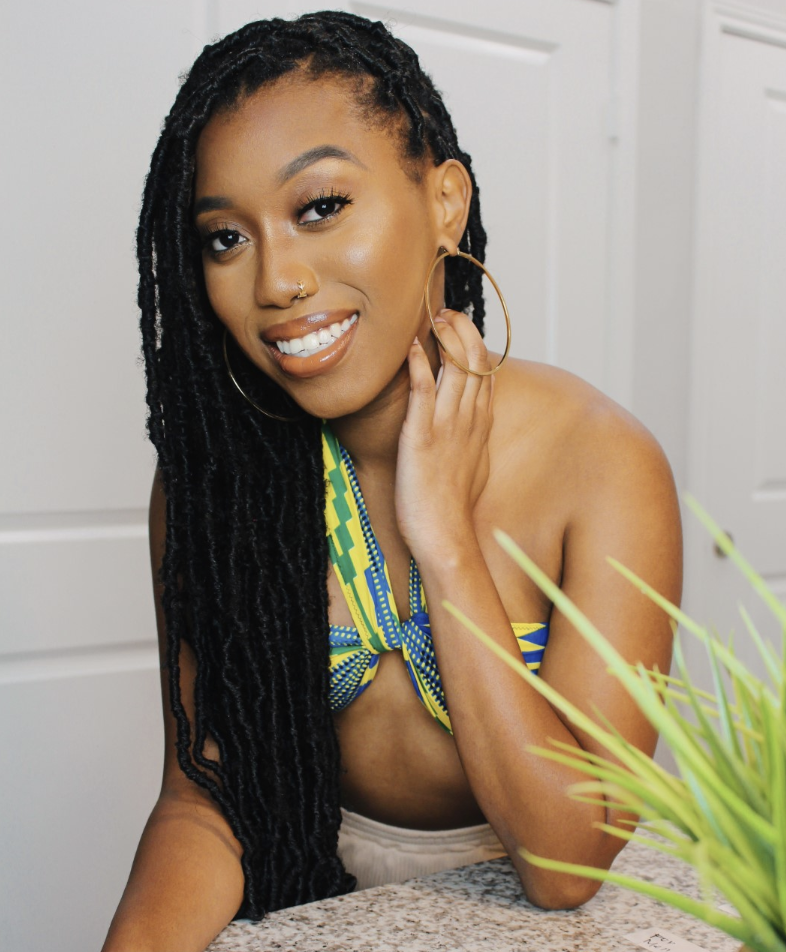 Photo of Alexa posing with elbow on table with plant. Dark brown complexion, dreadlocked hair, hoop earrings and has nose ring. Wearing a yellow and blue top and beige bottoms.