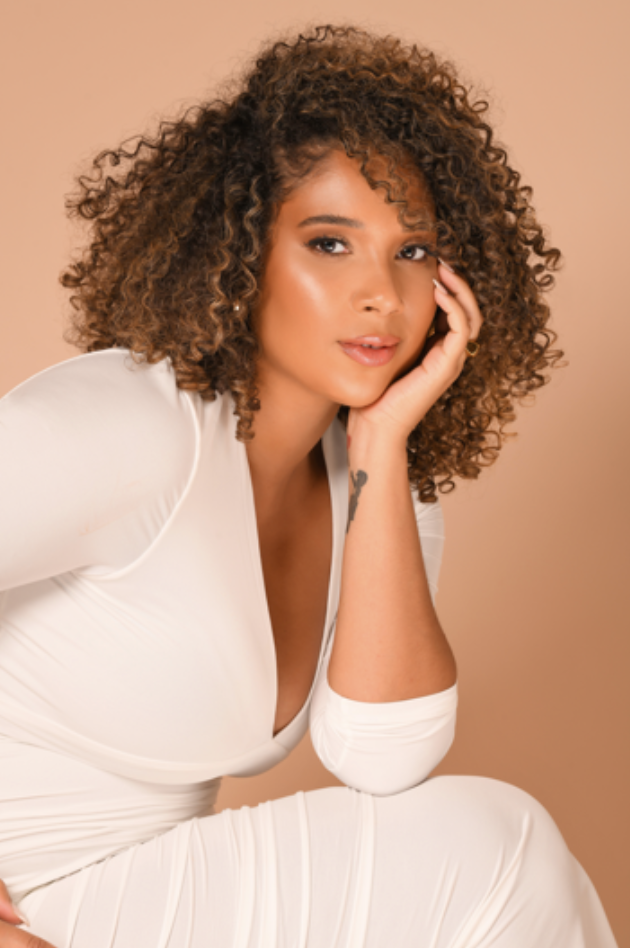 Photo of Ada posing with a brown background. She has brunette curly hair, brown complexion and is wearing lipstick. Wearing white outfit and has hand placed by her face.