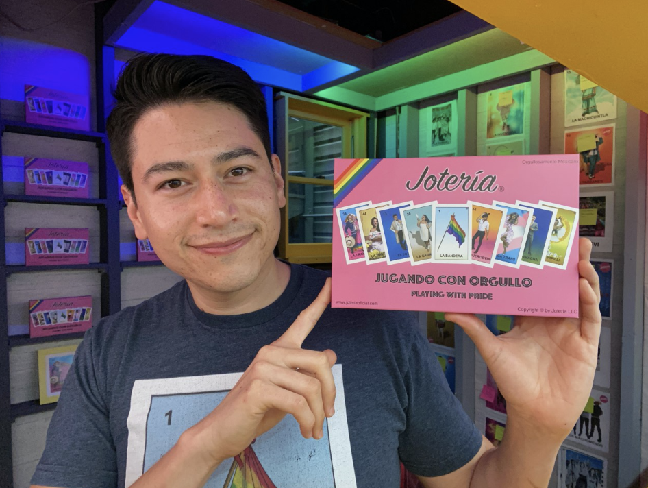 Photo of Antonio holding a pink box which says "Joteria, playing with pride." He is wearing a loteria shirt that is blue colored. Has white complexion and black combover hairstyle.