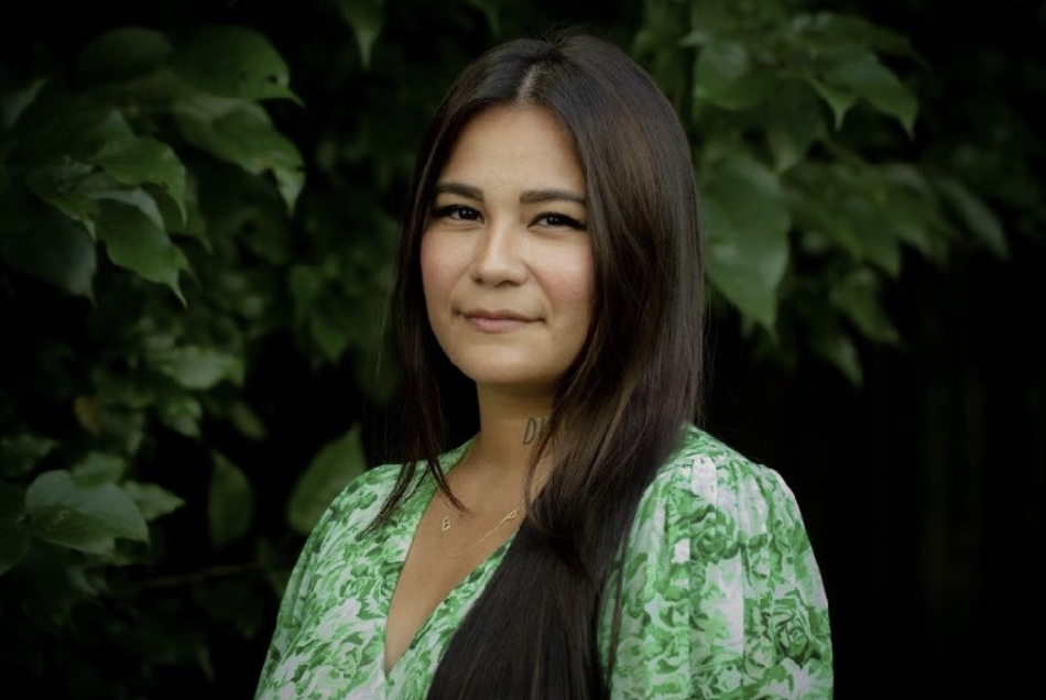 Picture of Prisca posing in front of green nature. Wearing a green and white top. Has fair complexion and rose pink cheeks and dark brown/black straight hair.