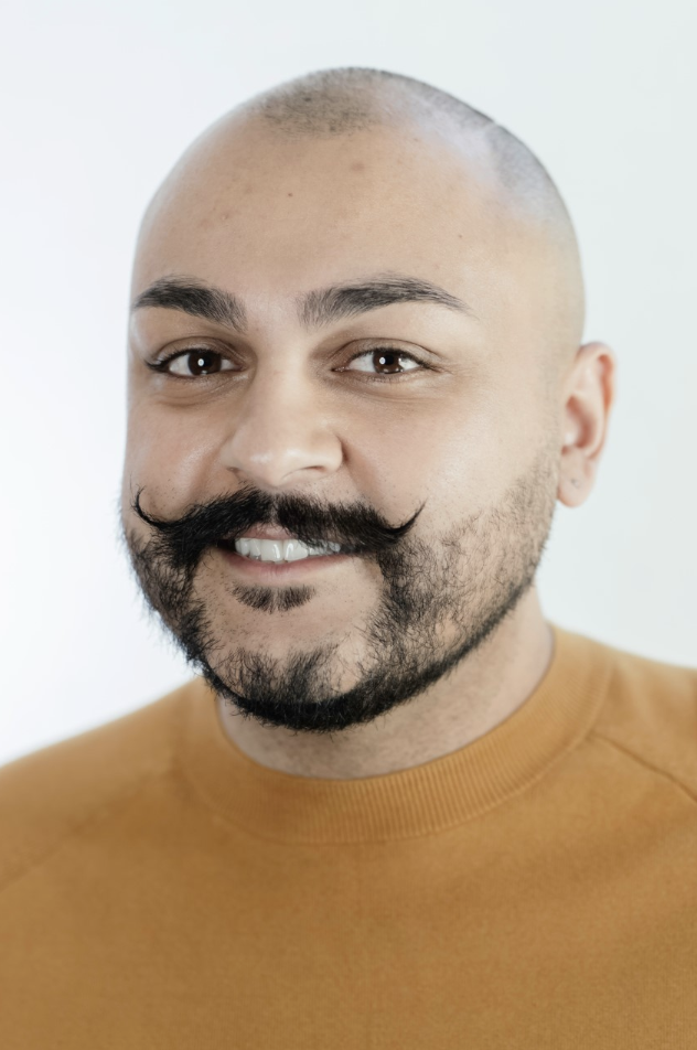 Photo of Sergio. Posing with white background, has shaved head, beard and curly mustache and is wearing brown top.