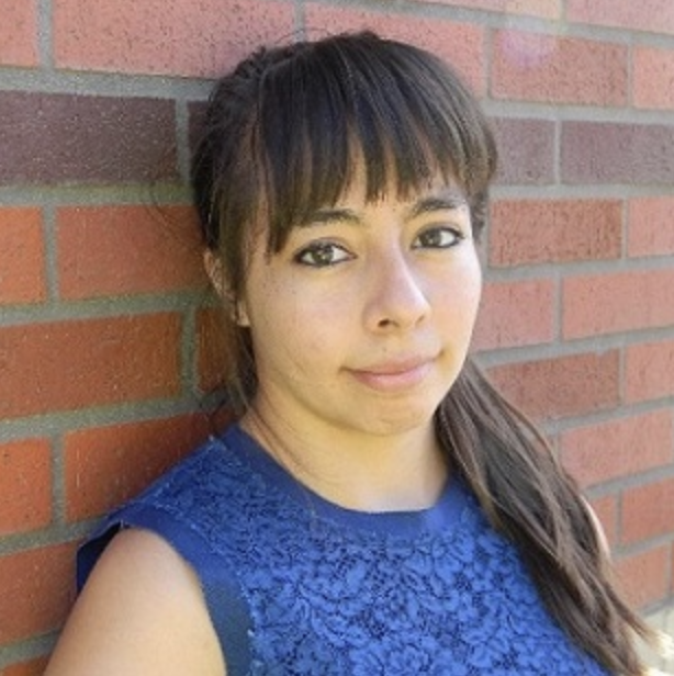 Picture of Blanca posing along a brick wall. Has brown skin, long brown straight hair with bangs, and is wearing a blue top.