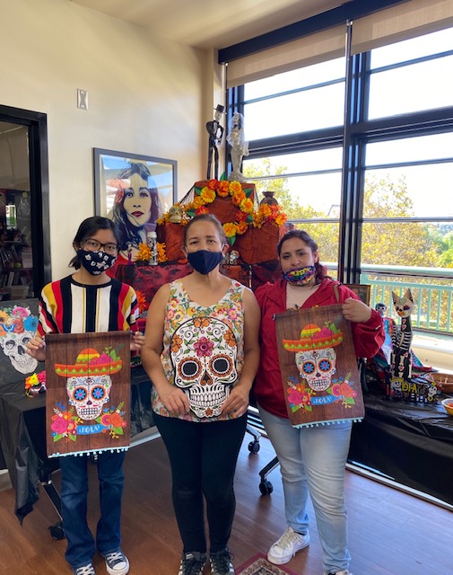 Three students posing in front of the altar holding sugar skull banners