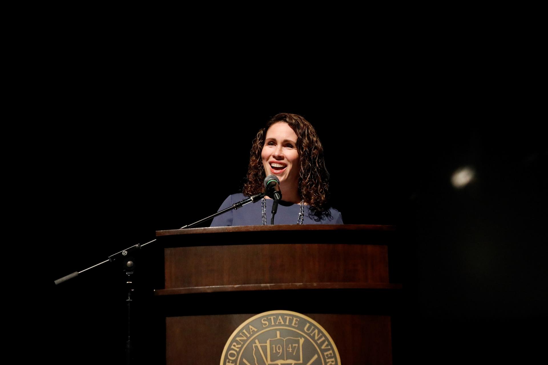 Tara Yosso speaking on a podium with a spotlight on her. She is wearing a blue top and has currly brown hair and beige skin tone. She is smiling.